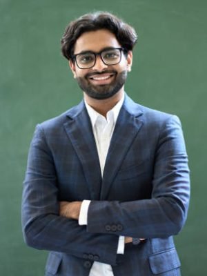 Smiling young indian business man professional manager wearing suit looking at camera standing arms crossed in office. Arab teacher or professor posing for portrait at work desk in front of blackboard